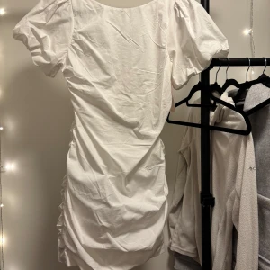 Vit kort klänning med puffärmar - Säljer en stilren vit kort klänning med puffiga kortärmar och rynkdetaljer. Klänningen har en elegant passform och en djupare rygg. aldrig använd, prislappen kvar. nypris 499:-