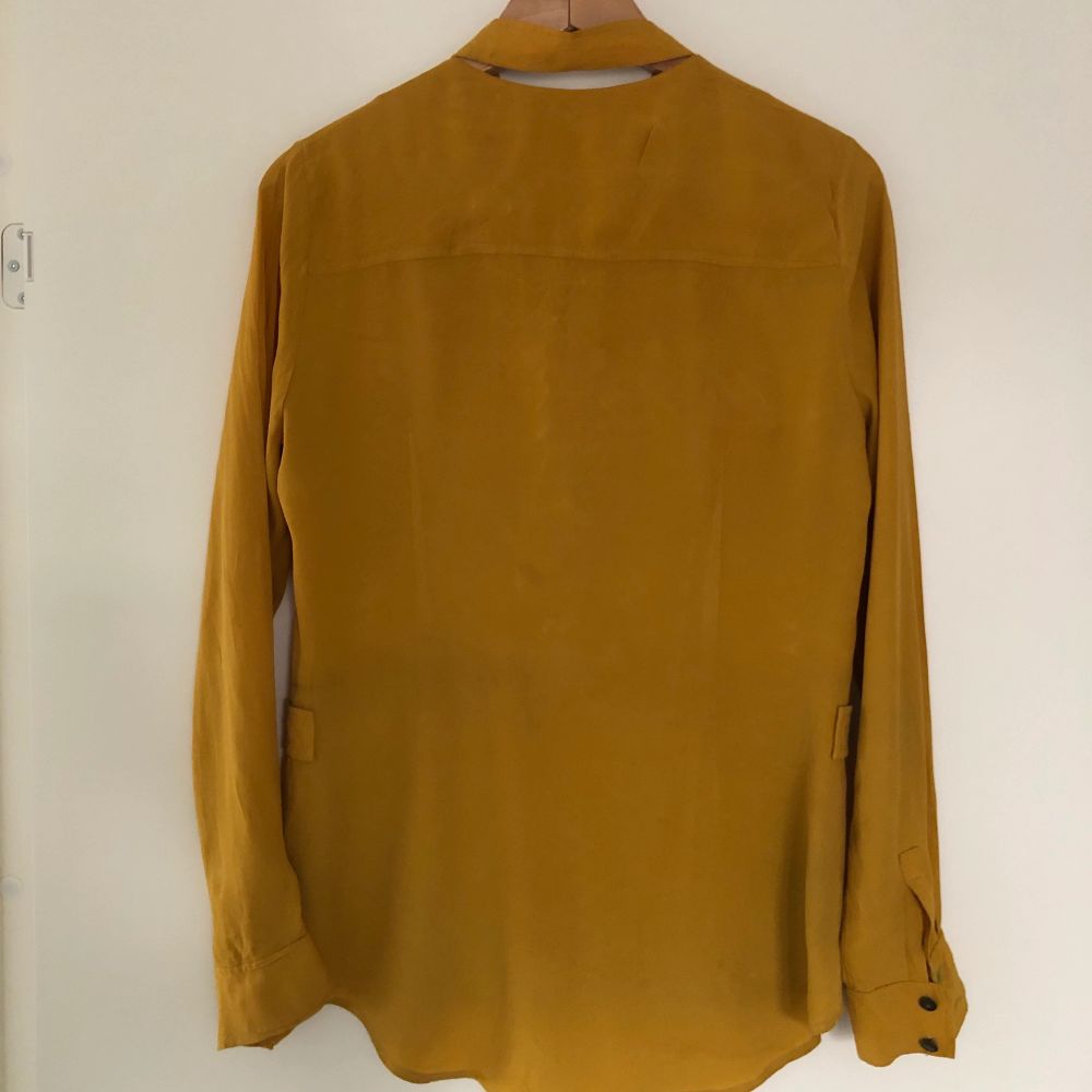 Beautiful blouse in mustard yellow colour. Front features a pleated apron-like detail and hidden button style. Back features split neckline. Fabric shows some signs of aging including decolorization and stains (e.g. around buttons). Skjortor.