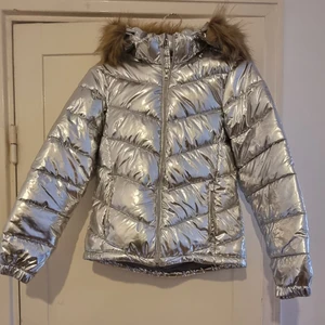 Mckinley jacka i silver med päls - Jacka i vind- och vattenavvisande material, PFC fri vattenavvisning. Avtagbar fodrad huva med avtagbar pälskant. Justerbar nederkant med dold dragsko. Fickor fram med dragkedja. Resår vid ärmslut. Reflexdetaljer bak, på ärm och fram. Vinterjacka, skidjacka. Storlek xs/160