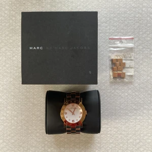Marc Jacobs klocka i roséguld - Armbandsur från Marc Jacobs i roséguld. Guldet är nött på baksidan (se bild 3). Jag mäter den inre omkretsen till ca 15cm, men det medföljer länkar man kan förlänga bandet med. OBS klockan behöver nytt batteri. Orginalkartong medföljer.