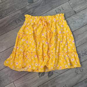Gul/orange kjol med blommigt mönster och volanger. Färgen är mer orange i verkligheter.