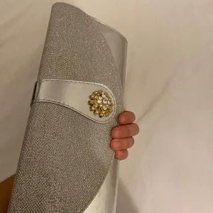 Silvrig handväska - Denna väskan är så snygg å elegant när man ska på t.ex bröllop eller bal. Du får plats med mobilen, läppglans (TJEJER DE VIKTIGT!!!)å annat. Har använts ungefär 2 gånger. (Köpare står för frakten)