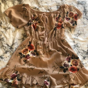 Somrig playsuit  - En somrig rosa playsuit med blommor på. Märket är Parisian. Köpt för några år sedan men knappt använd. Volanger vid slutet av shorts delen. Band vid kragen. Luftigt material.  Frågor?  Skriv i DM  Köparen står för frakten. 