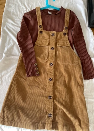 Hängselklänning  - Säljer en jätte fin hängselklänning och en  ribbad brun tröja med knappar i ärmen. Klänningen passar st 146-164.Pris-80kr Tröjan 146-164. Pris -50kr  