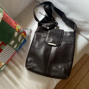 Axelremsväska i skinn  - Axelremsväska i mörkt läder, okänt märke. Slitage främst synligt på hörnen. Två fack inuti väskan, fläckfritt foder. Ca 30 x 25 cm. 