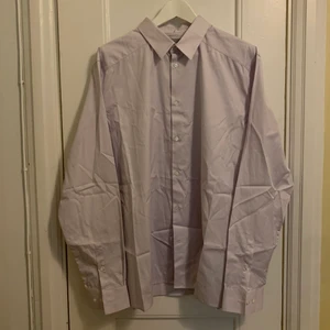 COS oversized skjorta - Klassisk herrskjorta från COS i en mycket svag ton av lila. Aldrig använd, därav nedvikt länge och ej struken. Fin uppkavlad till en ärmlös klänning, till cykelbyxor eller halvt instoppad till jeans. 100% bomullspoplin. Köpte den för färgen!