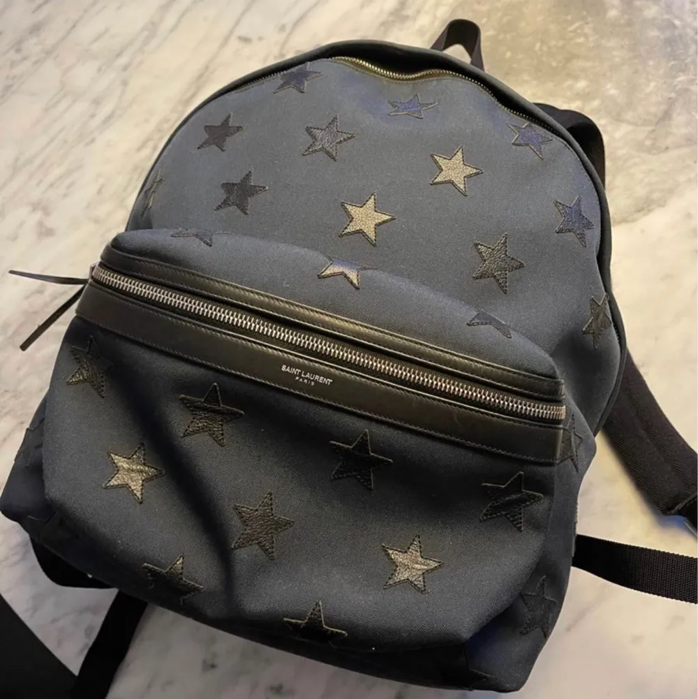 ryggsäck från saint Laurent i mörkblått med star motivet.  Bra väska som rymmer det mesta, laptop/iPad fack, nyckelhållare. Nypris 8995 Knappt använd, endast väskan säljs. Väskor.