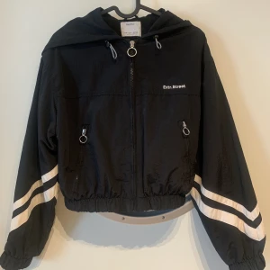 Cropped Windbreaker - Bershka cropped windbreaker jacka med vita streck på ärmarna med luva och två fickor. 