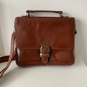 Axelremsväska / portfölj i läder - Så fin axelremsväska / portfölj i läder. Axelremmen är justerbar men går även att ta bort om man vill💼I väldigt gott skick!🌟 Väskans mått (Bredd x höjd): 23x19 cm