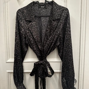 Leopardmönstrad blus med knytning - Grå blus från BikBok med svart leopardmönster. Saknas en knapp på en av armarna men är två knappar på varje arm så det syns inte tydligt. Har katt i hemmet men ej där kläderna är.