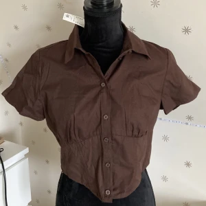 Brun topp - Oanvänd brun blus med krage och korta ärmar. Extra knapp medföljer. Säljs då den är lite stor för mig