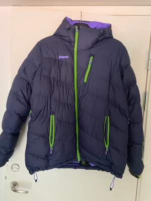 Fantastisk varm dunjacka, perfekt för vintern - Mörkblå varm dunjacka med lila insida och snygga gröna detaljer från Bergans of Norway säljes i nyskick, använd ca 2 ggr, då den tyvärr var för stor. Nypris: 3999