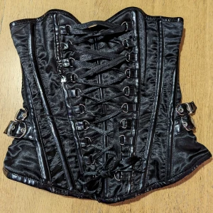 Burleska overbust korsett - En punkig overbust korsett från Burleska i storlek M. Lackdetaljer på skenor och spännen på sidorna och svart satin som bas.  Fint skick utan några defekter.  Mått från tillverkaren:  Bust: 84cm  Waist: 76cm  Eu size: 38-40