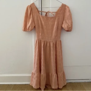 Orange klänning  - Ljusorange klänning med öppen rygg, puffärmar, square neck, knytning i midjan och rosett i ryggen, volanger längst ner i bäckebölja. Stretchig och SÅ bekväm - den perfekta modellen. 