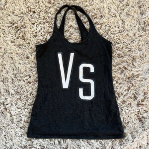 Mörkgrå VS linne från Victoria's Secret - Snygg mörkgrå linne från Victoria's Secret med stora vita VS-bokstäver framtill. Toppen har dubbla smala axelband och ärmlös design, perfekt för träning eller chill. Mjuk bomullsblandning som känns skön mot huden. Tyvärr har linnet ett håll vid högra bröstet. Storlek M Från Victoria secret