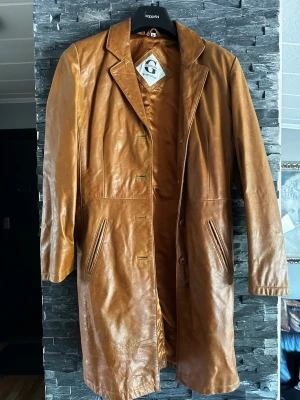 Lång cognac färgad skinnkappa med klassisk krage - Snygg lång cognac färgad skinnkappa med klassisk krage och knäppning framtill. Jackan har två snedställda fickor och en stilren design som ger en retrovibe. Perfekt för dig som vill sticka ut med en tidlös look.