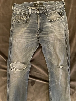 Replay jeans  - Säljer ett par blå jeans från Replay med slitningar och hål på vänster knä. Jeansen har klassisk femficksdesign, ljus tvätt och kontrastsömmar. Perfekta för en avslappnad och trendig look.