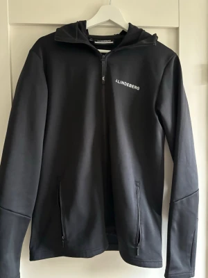 Svart hoodie från J.Lindeberg - Snygg svart hoodie från J.Lindeberg med dragkedja framtill och två fickor med dragkedja. Märkets logga syns på bröstet och huvan har en diskret logodetalj. Tillverkad i ett mjukt och stretchigt material som passar perfekt till vardagslooken.