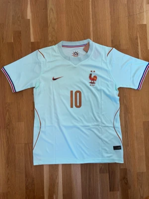 Frankrike fotbollströja Mbappé #10 - Snygg ljusblå Frankrike fotbollströja från Nike med Mbappé och nummer 10 på ryggen. Tröjan har korta ärmar, broderad tupp och två stjärnor på bröstet samt röda och blå detaljer på ärmsluten. Perfekt för match eller träning!