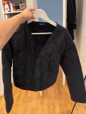 Svart quiltad cardiganjacka - Snygg svart quiltad cardiganjacka med knappar framtill och två stora fickor. Jackan har en avslappnad passform och V-ringning, perfekt att slänga över en topp. Materialet är vadderat och ger en trendig look.