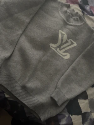 Grå sweatshirt med vit LV-logga - Snygg grå sweatshirt med stor vit LV-logga på bröstet. Tröjan har rund halsringning, ribbade muddar vid ärmslut och nederkant samt en avslappnad passform. Perfekt för en casual och trendig look.