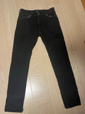 Svarta Jack&Jones slim fit jeans (28/32) - Säljer ett par svarta slim fit jeans med fem fickor och vita kontrastsömmar. Jeansen har klassisk knappgylf och raka ben. Perfekta till en avslappnad stil och passar till det mesta i garderoben.