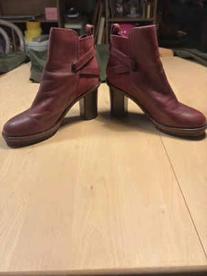 Vinröda stövletter med klack  - Snygga vinröda stövletter i skinn med rundad tå och kraftig klack. Dekorativa sömmar och remdetaljer runt ankeln ger en cool look. Perfekta för dig som vill ha både stil och höjd på steget.