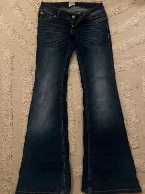 Mörkblå bootcut jeans låg midja  - Säljer ett par mörkblå bootcut jeans med snygga slitningar och klassiska fem fickor. Jeansen har låg midja och vida ben, perfekta för en retroinspirerad look. Tillverkade i stretchigt denimtyg för skön passform.