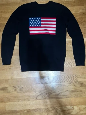 Svart stickad tröja med USA-flagga - Svart stickad tröja med en stor amerikansk flagga i rött, vitt och blått på bröstet. Tröjan har rund halsringning och långa ärmar. Ribbstickade muddar vid ärmslut och nederkant. Perfekt för dig som gillar statement-plagg.