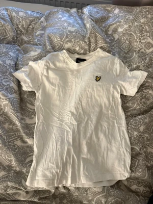 Vit t-shirt från Lyle & Scott - Säljer en vit t-shirt från Lyle & Scott med klassisk gul logga på bröstet. T-shirten har korta ärmar, v-ringning och är gjord i mjuk bomull. Enkel och stilren design som passar till det mesta.