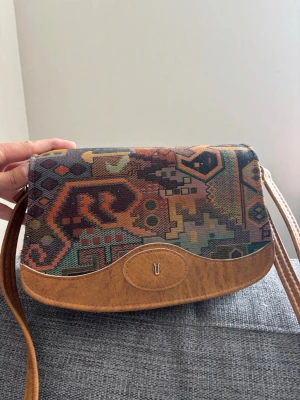 Mönstrad axelväska i retrostil - Unik axelväska med färgglatt geometriskt mönster i tyg och detaljer i brunt konstläder. Väskan har justerbar axelrem och en liten metallogo framtill. Perfekt för dig som gillar vintagevibbar och vill sticka ut med din stil. Mått: 25x17x10 cm  Superfint skick förutom något ”naggad” på kanten se bild men den har hållit sig riktigt fin med tanke på tillverkad 90-tal