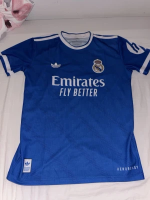 Blå Real Madrid matchtröja Adidas - Snygg blå Real Madrid matchtröja från Adidas med vita detaljer och klubbmärke på bröstet. Tröjan har 'Emirates Fly Better' tryckt framtill och är tillverkad i lätt och ventilerande AEROREADY-material. Perfekt för fotbollsträning eller att bära till vardags.