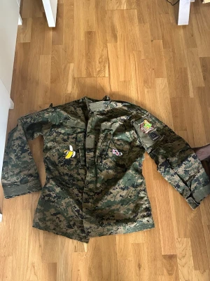 Kameramönstrad militärjacka med patches - Cool militärjacka med digitalt kamouflagemönster i gröna och bruna nyanser. Jackan har flera patches, bland annat Top Gun, Slovakien och roliga motiv som banan och panda. Praktiska fickor framtill och kardborre vid halsen. Perfekt för dig som gillar army style.