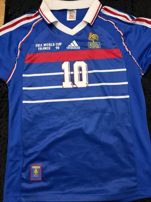 Frankrike VM 98 tröja Adidas blå - Säljer en ikonisk blå Frankrike fotbollströja från VM 1998 med nummer 10 på bröstet. Tröjan har vita och röda detaljer, klassisk krage och officiella FIFA World Cup och Adidas-loggor. Perfekt för dig som älskar retro fotbollsstil.