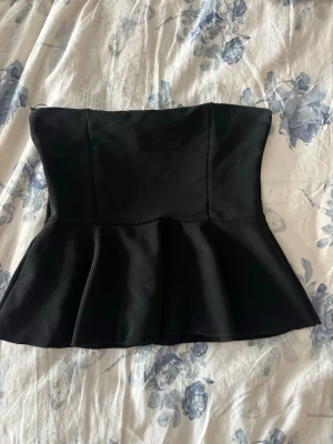 Svart peplum topp axelbandslös - Snygg svart axelbandslös peplum topp med markerad midja och utsvängd nederdel. Toppen har en stilren och elegant design, perfekt att matcha med jeans eller kjol. 