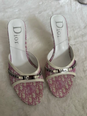 Rosa Dior pumps med silverdetaljer - Säljer ett par rosa och vita Dior pumps med ikoniskt monogrammönster. Skorna har öppen tå, smal klack och silverfärgad kedjedetalj med Dior-logga framtill. perfekta för dig som vill sticka ut.  Står storlek 40, passar 39