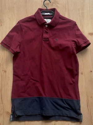 Vintage piké från Abercrombie & Fitch - Snygg vinröd pikétröja från Abercrombie & Fitch med klassisk krage, två knappar och mörkblå detalj längst ner. Liten broderad logga på bröstet. Perfekt till jeans eller shorts för en avslappnad look.
