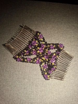 Hårkam med lila och svarta pärlor - Dubbel hårkam dekorerad med lila och svarta pärlor samt små gula detaljer. Metallkammarna binds ihop av elastiska pärlband i ett korsande mönster. Perfekt för att skapa en unik frisyr med extra bling.