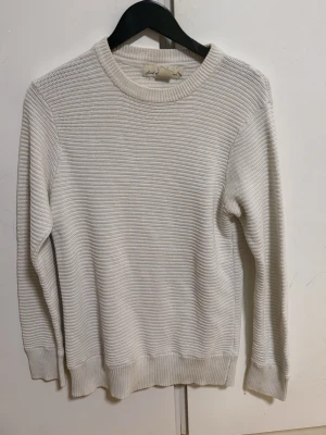 Vit ribbstickad tröja - Säljer en stilren vit ribbstickad tröja med rund halsringning och långa ärmar. Tröjan har breda muddar vid ärmslut och nederkant, vilket ger en klassisk och enkel look. Perfekt att matcha med jeans eller kjol.