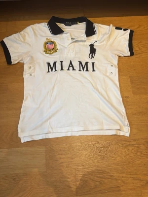 Polo Ralph Lauren Miami 5 pike - Vit pikétröja från Polo Ralph Lauren med svart krage och ärmslut. Stort MIAMI-tryck och logga framtill, samt siffran 5 på ryggen. Broderad detalj på bröstet och klassisk pikéknäppning. Perfekt för en sportig och stilren look.