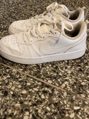 Vita Nike sneakers i läder - Snygga helvita sneakers från Nike med klassisk låg siluett. Skorna har perforerad tå, vita skosnören och diskret Nike-logga på sidan. Tillverkade i läder med bekväm vadderad insida och slitstark yttersula.