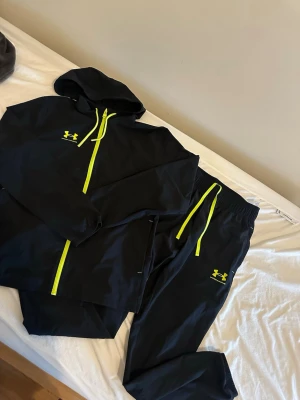 Under Armour Tracksuit  - Snyggt svart träningsset från Under Armour med matchande hoodie och byxor. Setet har neongula detaljer som dragkedja, snören och logga. Perfekt för gymmet eller löprundan.