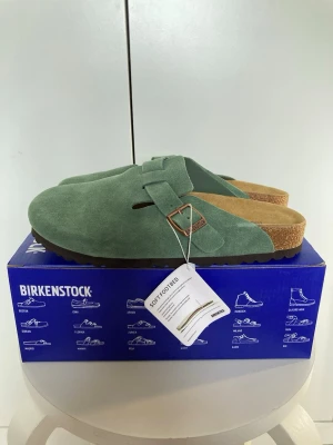 Gröna Birkenstock tofflor mocka - Säljer ett par gröna Birkenstock tofflor i mjuk mocka med justerbart spänne i metall. Klassisk slip-in modell med korksula och ergonomisk fotbädd för extra komfort. Perfekta för dig som vill ha både stil och bekvämlighet.