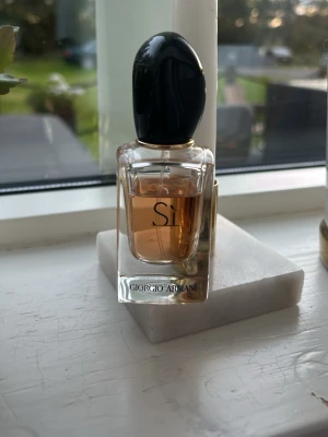 Giorgio Armani Sì parfym - Elegant parfymflaska från Giorgio Armani med namnet Sì. Flaskan är genomskinlig med en svart, glansig kork och innehåller en bärnstensfärgad vätska. Klassisk och stilren design som passar perfekt på parfymhyllan.