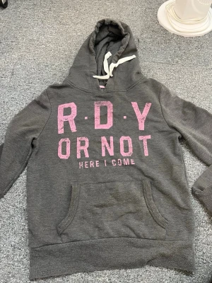 Grå hoodie med rosa text och huva - Mysig grå hoodie med stor rosa text 'R.D.Y OR NOT HERE I COME' på bröstet. Tröjan har en klassisk känguruficka framtill och vit dragsko i huvan. Perfekt för chill dagar eller när du vill sticka ut lite extra. Tillverkad i mjukt sweatshirtmaterial.