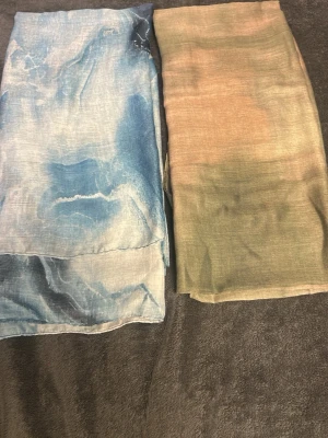 Två tunna sjalar i blått och beige - Säljer två sjalar i tunt och lätt material, en med blå och vita nyanser och en med beige och gröna toner. Båda har ett mjukt, abstrakt mönster och är perfekta att slänga över axlarna för en cool look. De är helt nya ✨