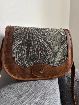 Brun axelväska med paisleymönster - Snygg axelväska i brunt konstläder med lock i tyg med paisleymönster i svart, grått och beige. Väskan har en dekorativ liten metallbricka med kvarnmotiv på framsidan och justerbar axelrem. Perfekt för dig som gillar vintagevibbar och unika detaljer. Mått: 25x18x10 cm