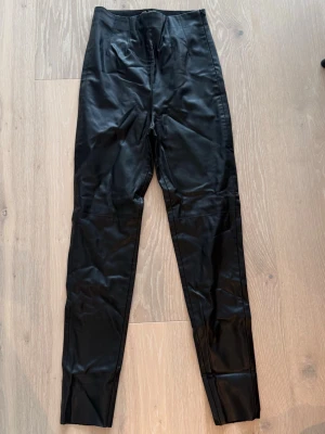 Svarta fuskskinnleggings - Snygga svarta leggings i fuskskinn med hög midja och smal passform. Materialet ger en cool och trendig look som passar perfekt till en streetstyle-outfit. Byxorna har inga synliga fickor eller dragkedjor för en clean känsla.