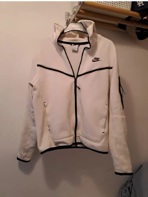 Vit hoodie med dragkedja från Nike - Snygg vit hoodie från Nike med svart dragkedja och svarta detaljer. Hoodien har två fickor framtill, en huva och en liten ficka på ärmen. Tillverkad i mjukt material som känns skönt mot huden. Perfekt för en sportig och avslappnad stil.