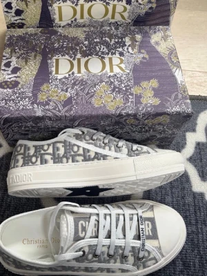 Grå Dior sneakers med monogram - Snygga låga sneakers från Dior i grått och vitt med klassiskt Dior-monogram över hela skon. Skorna har vita snören, vit sula och detaljer med texten 'Christian Dior' på sidan och hälen. Tillverkade i textil och gummi för en modern och exklusiv look.